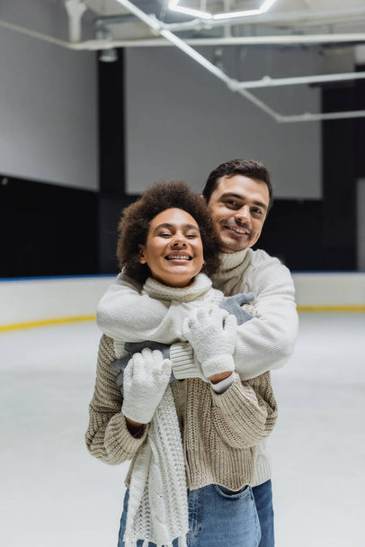 Χαρούμενος άντρας αγκαλιάζει την Αφροαμερικανή φιλενάδα του και κοιτάζει την κάμερα στο παγοδρόμιο  - Φωτογραφία, εικόνα