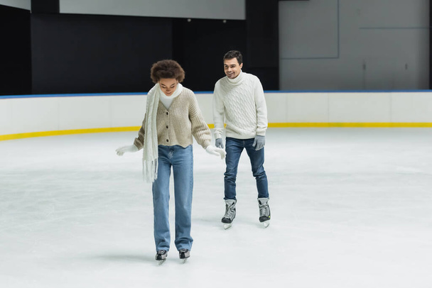 Donna afroamericana in sciarpa e guanti pattinaggio su ghiaccio vicino fidanzato sulla pista di pattinaggio  - Foto, immagini