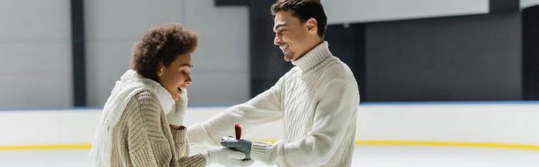 Vista lateral do homem sorridente em suéter dando anel proposta para a namorada afro-americana no ringue de gelo, banner  - Foto, Imagem