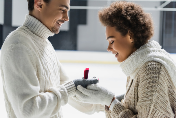 Vue latérale de l'homme en pull tenant la bague de fiançailles près de la petite amie afro-américaine sur la patinoire  - Photo, image
