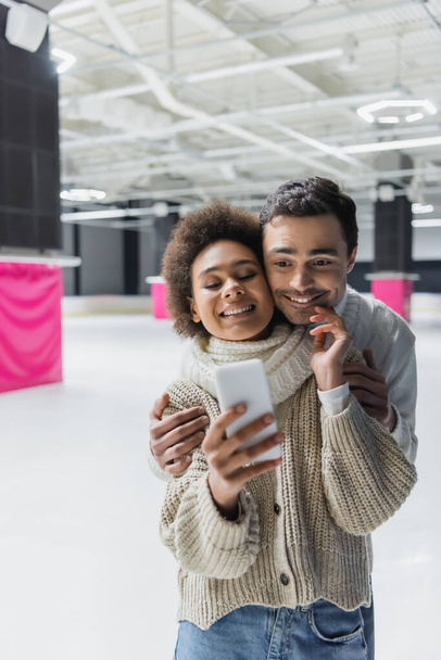 Sonriente mujer afroamericana tocando novio y tomando selfie en smartphone en pista de hielo  - Foto, Imagen