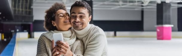 Man hugging african american girlfriend with cellphone on ice rink, banner  - Photo, Image