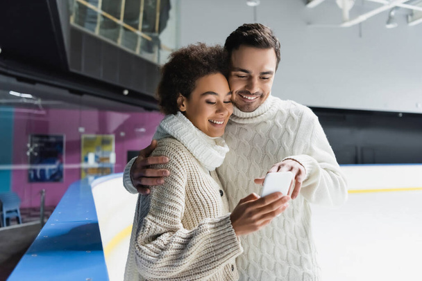 Lächelnder Mann umarmt afrikanisch-amerikanische Freundin und benutzt Smartphone auf Eisbahn  - Foto, Bild