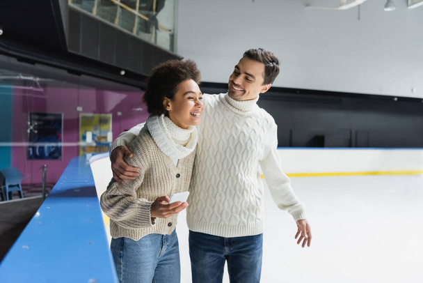 Positive man in sweater hugging african american girlfriend with smartphone on ice rink  - Φωτογραφία, εικόνα
