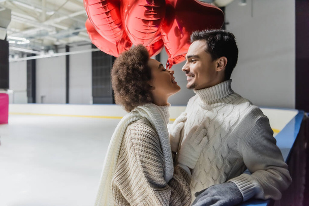 Vista lateral da mulher americana africana sorridente olhando para o namorado perto de balões em forma de coração na pista de gelo  - Foto, Imagem
