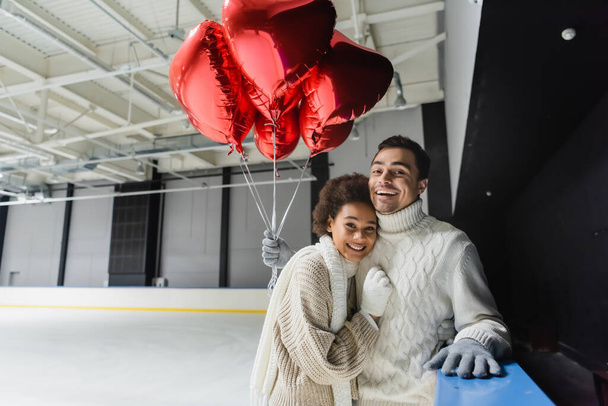 Cheerful interracial couple holding heart shaped balloons and looking at camera on ice rink  - Foto, Bild