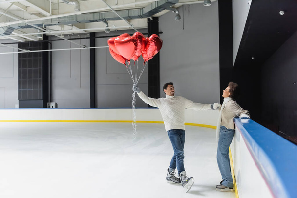Cheerful african american woman holding hand of boyfriend with heart shaped balloons on ice rink  - Fotoğraf, Görsel