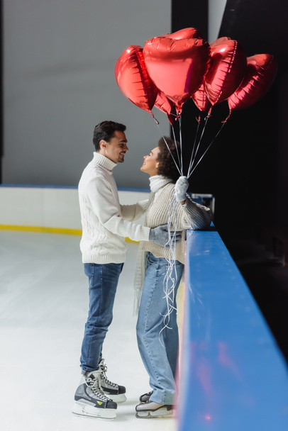 zijaanzicht van jongeman in schaatsen knuffelen afrikaanse amerikaanse vriendin holding hartvormige ballonnen op ijsbaan  - Foto, afbeelding