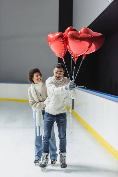Positive multiethnic couple with heart shaped balloons ice skating on rink  - Φωτογραφία, εικόνα