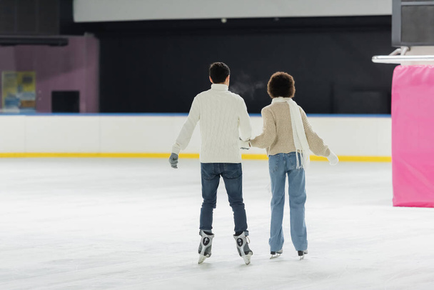 Vista trasera de la pareja interracial patinaje sobre hielo juntos en la pista de patinaje  - Foto, Imagen
