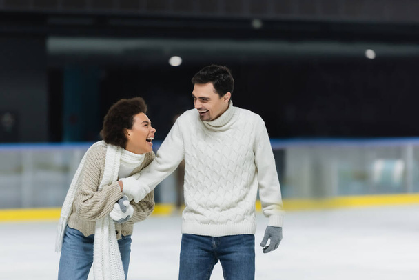 Cheerful african american woman holding hand of boyfriend in sweater and gloves on ice rink  - Φωτογραφία, εικόνα