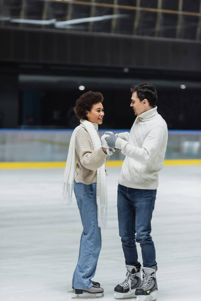Jeune homme en pull tenant la main de la petite amie afro-américaine en gants sur la patinoire  - Photo, image