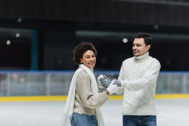 Cheerful african american woman looking at camera and holding hands of boyfriend on ice rink  - Foto, Imagen