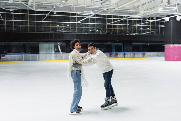 Positive multiethnic couple in gloves looking at camera while having fun on ice rink  - Fotoğraf, Görsel