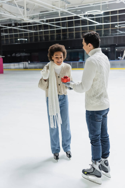 Smiling african american woman looking at heart shaped gift near boyfriend on ice rink  - Фото, зображення