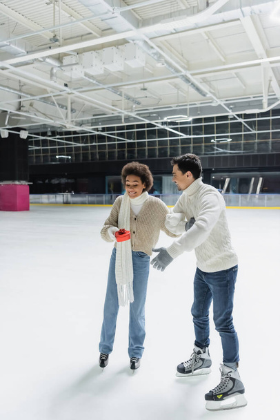 Excited african american woman holding heart shaped box near boyfriend on ice rink  - Фото, изображение