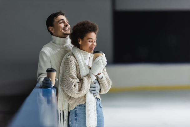 Positive interracial couple in gloves holding coffee to go on ice rink  - Photo, Image