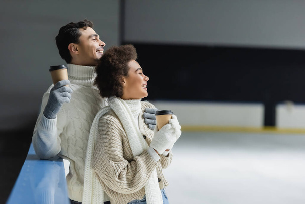 Side view of cheerful interracial couple holding coffee to go and looking away on ice rink  - Photo, Image