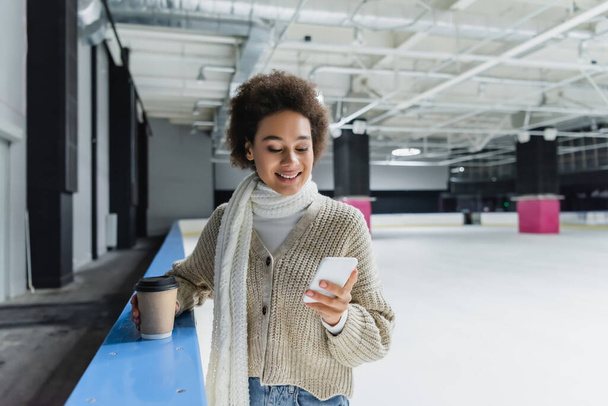 glimlachen Afrikaans amerikaanse vrouw in sjaal met behulp van smartphone en het houden van koffie te gaan op de ijsbaan  - Foto, afbeelding