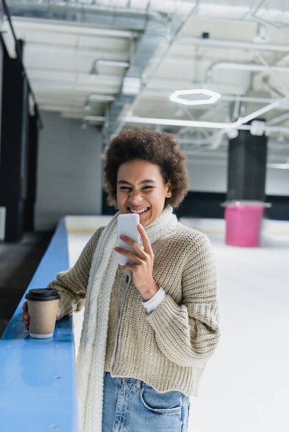 Mosolygó afro-amerikai nő gazdaság smartphone és papír csésze a jégpályán  - Fotó, kép