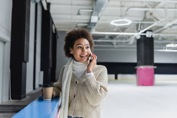 Positive african american woman talking on smartphone and holding paper cup on ice rink  - Foto, Bild