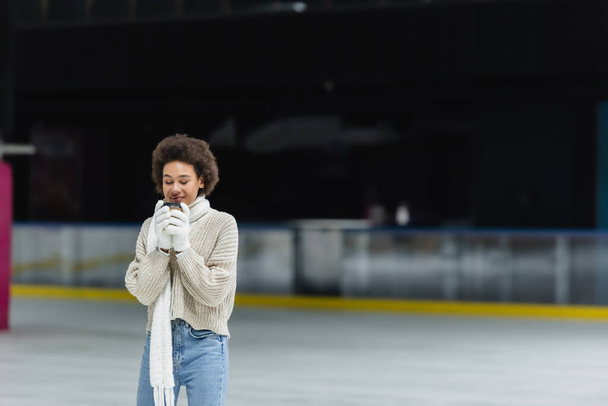 Junge Afroamerikanerin in Handschuhen und Schal mit Kaffee zum Eislaufen  - Foto, Bild