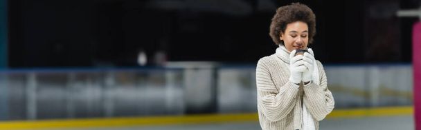 Pleased african american woman in warm clothes holding coffee to go on ice rink, banner  - Fotoğraf, Görsel