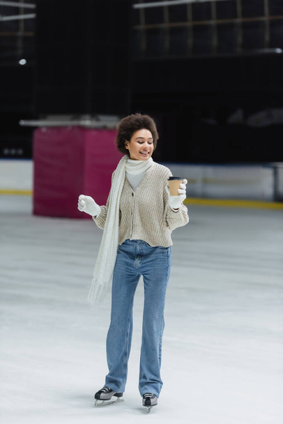 Smiling african american woman ice skating and looking at takeaway coffee in paper cup on ice rink  - Foto, Bild