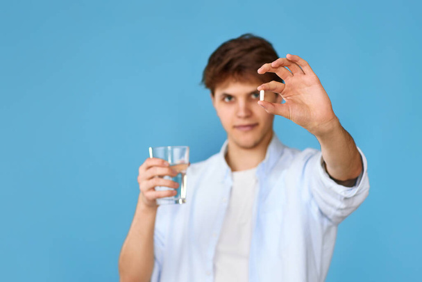 Young handsome man holding white tablet and glass of water on blue background. focus on pill - Foto, afbeelding