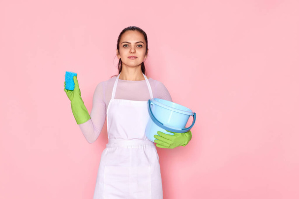 young confident woman in rubber gloves and cleaner apron cleaning with blue sponge and bucket on pink background. - Foto, afbeelding