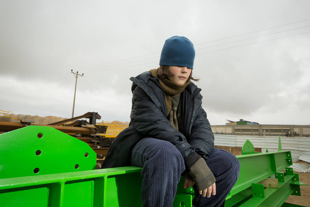 cool stylish caucasian boy with the blue hat on his face covering his eyes in a coat and hat with a scarf posingon green metal structures in an industrial area against a cloudy greysky on a rainy day - Fotó, kép