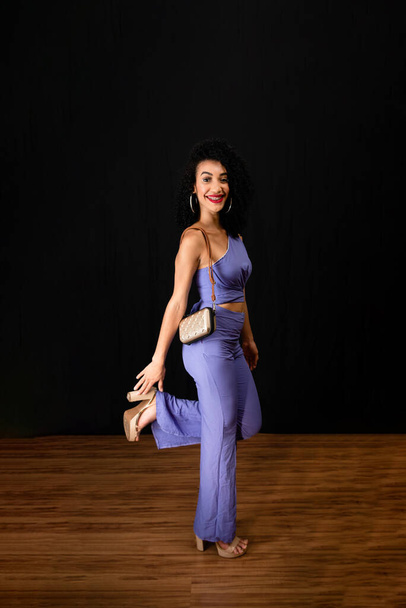 Portrait of young beautiful, joyful woman standing in studio. Holding a bag. wearing blue clothes. Against black background. - Foto, afbeelding