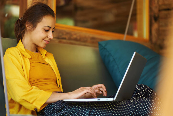 Πορτρέτο νεαρή γυναίκα στο καφέ με το laptop της στα γόνατα πληκτρολογώντας κάτι, γράφοντας νέα θέση στο blog της. Καλοκαιρινές διακοπές και μερικές φορές εργάζονται εξ αποστάσεως - Φωτογραφία, εικόνα