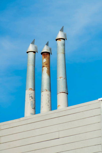 Gray metal chimney pipes with some rust and wooden slatted building exterior facade with blue sky background. Some clouds in the midday sun on the side of the building or structure. - Foto, Imagen