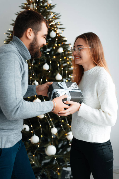 A young man and a young girl loving each other hug and kiss in cozy sweaters near a small Christmas tree. - Foto, Imagen