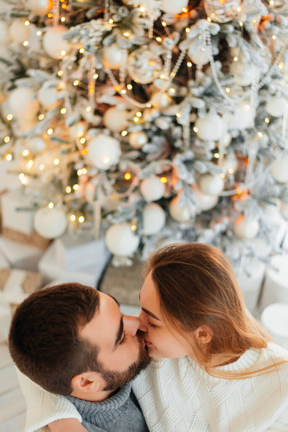 A young man and a young girl loving each other hug and kiss in cozy sweaters near a small Christmas tree. - Foto, afbeelding