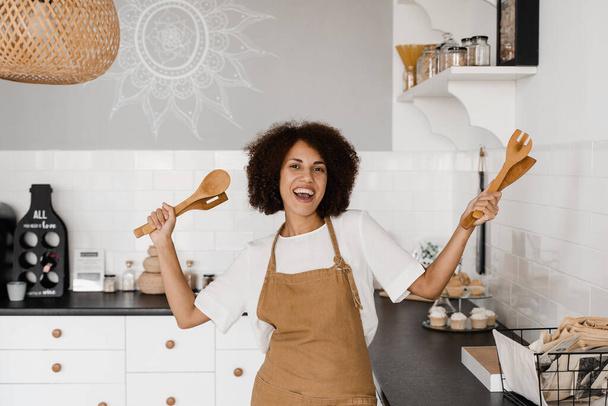 Afro-Amerikalı aşçı kız spatulayla yemek pişirmek ve eğlenmek için dans ediyor. Önlüklü mutlu Afrikalı ev kadını kahvaltı hazırlamadan önce mutfakta şarkı söyleyip dans ediyor. - Fotoğraf, Görsel