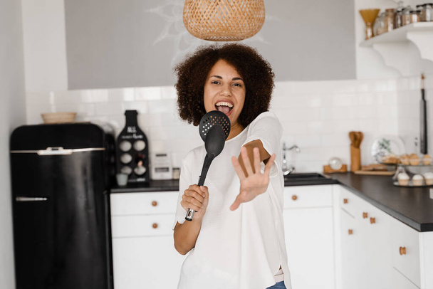 Joyeuse jeune femme africaine s'amusant avec des ustensiles de cuisine. Femme au foyer afro-américaine dans un tablier chantant avec des spatules pour la cuisson microphone sur la cuisine - Photo, image