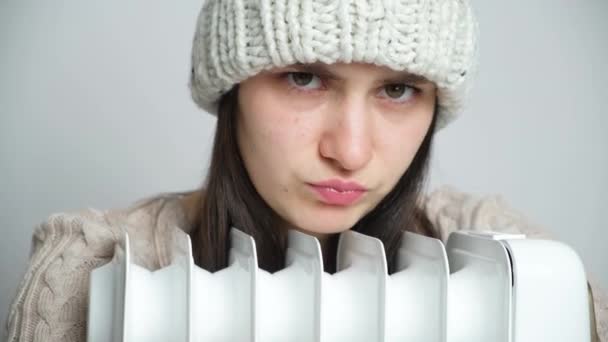 Egy téli gyapjú kalapos nő megölel egy elektromos olajmelegítőt, és szomorúan néz ki, lefagyasztva. - Felvétel, videó