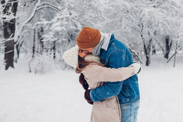 Young stylish loving couple kissing in winter park. Man and woman hugging while walking and enjoying snowy weather outdoors under falling snow. Valentine's day - Photo, Image