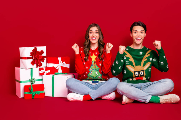 Фотографія двох чудових вражених людей, які сидять на підлозі, піднімають кулаки стопки новорічної подарункової коробки ізольовані на червоному тлі
. - Фото, зображення