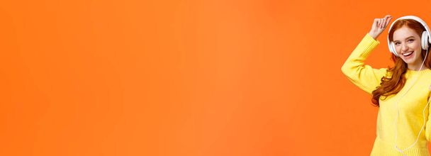 Dansons. Joyeux insouciant moderne hipster girl avec les cheveux bouclés rouges et les taches de rousseur, danse lever la main et tenant smartphone, écouter de la musique avec de nouveaux écouteurs en ligne commandés, fond orange
. - Photo, image