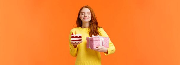 Oslava, lidé a b-denní koncept. Zasněný pozitivní a něžná zrzka žena slaví narozeniny, drží kus dortu se svíčkou a zavřít oči šťastný, držet roztomilý růžový zabalený dárek. - Fotografie, Obrázek
