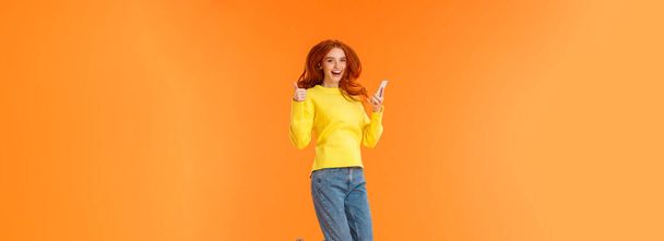 Vertical full-length shot carefree, excited and pleased good-looking ginger girl in sweater, jeans, jumping over orange background, holding smartphone show thumbs-up and smiling approvingly. - Photo, Image