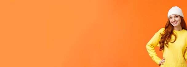 Menina moderna hipster jogando bolas de neve com amigos durante as férias de inverno, mostrando sinal de paz como posando foto perto boneco de neve, vestindo gorro branco e suéter sobre fundo laranja
. - Foto, Imagem