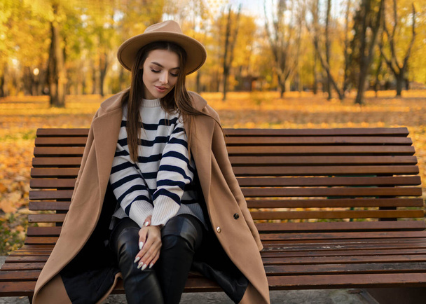 portrait d'une jolie femme souriante et élégante aux cheveux longs marchant dans le parc, vêtue d'un manteau marron chaud automne mode tendance, style de rue avec chapeau. - Photo, image