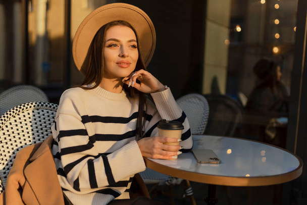 krásná usmívající se žena ve stylovém oblečení sedí u stolu v klobouku a svetru, romantická šťastná nálada, čeká na přítele na rande v kavárně, jaro-letní módní trend, pití kávy. - Fotografie, Obrázek