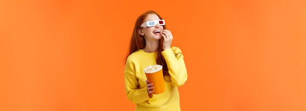 Felice e divertito rossa ragazza piace guardare film nei teatri, frequentare il cinema, mangiare popcorn e guardare lo schermo in bicchieri 3d, sorridente portato via film interessante, sfondo arancione
. - Foto, immagini