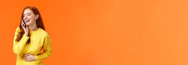 Kommunikation, Technologie und Menschen-Konzept. fröhliches hübsches rothaariges Hipstermädchen, das unbeschwert telefoniert und lacht, bauchnahe Augen, Smartphone in Ohrnähe, orangefarbener Hintergrund. - Foto, Bild