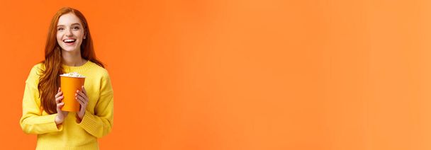 fröhlich niedlich rothaarige Lockenkopf Freundin hält Popcorn-Chat mit Freund vor Filmstart im Kino, lächelt fröhlich, wie bei Premieren im Kino, stehend überglücklich orange Hintergrund. - Foto, Bild